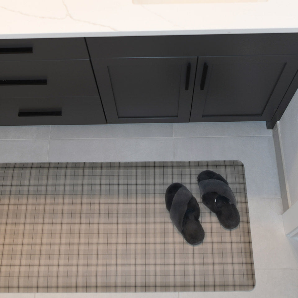 Overhead images of Brisbane Plaid anti-fatigue comfort mat in the color tan with a pair of slippers resting on top in a bathroom