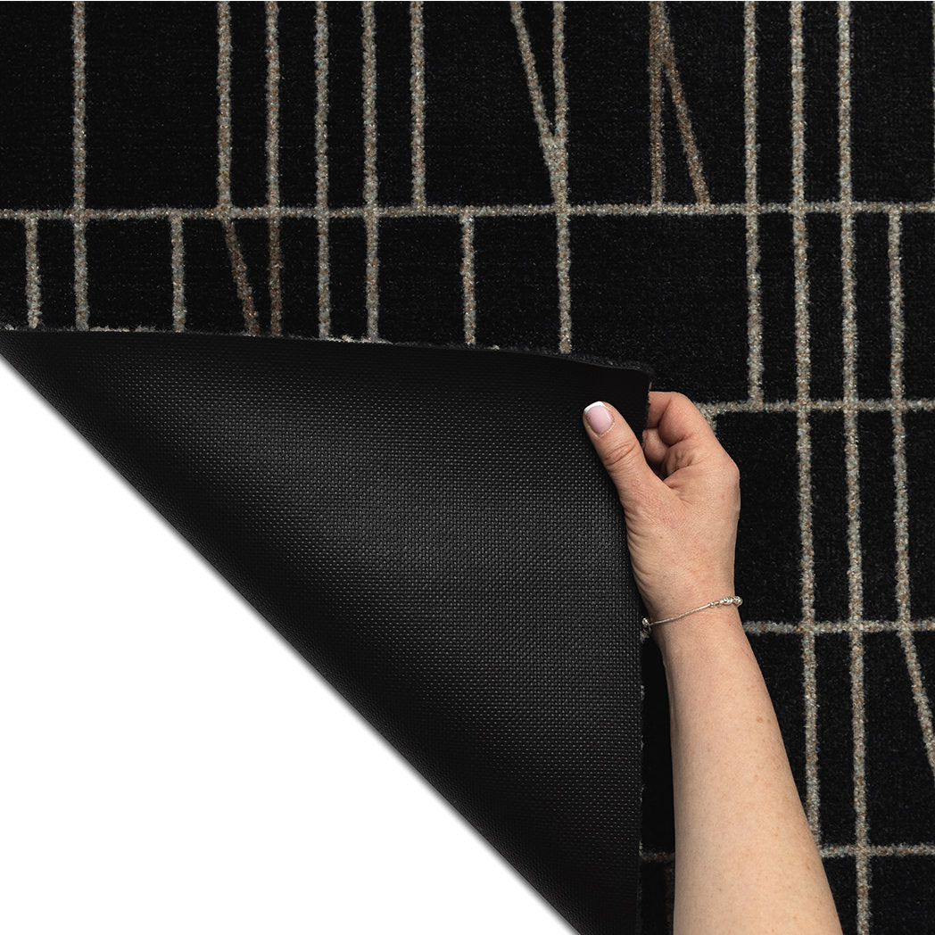 Hand lifting the corner of the Maxcent Biblioteca mat in charcoal to show the textured, slip-resistant rubber backing. The durable underside helps keep the mat securely in place, while the top features a modern grid pattern on a charcoal carpeted surface.