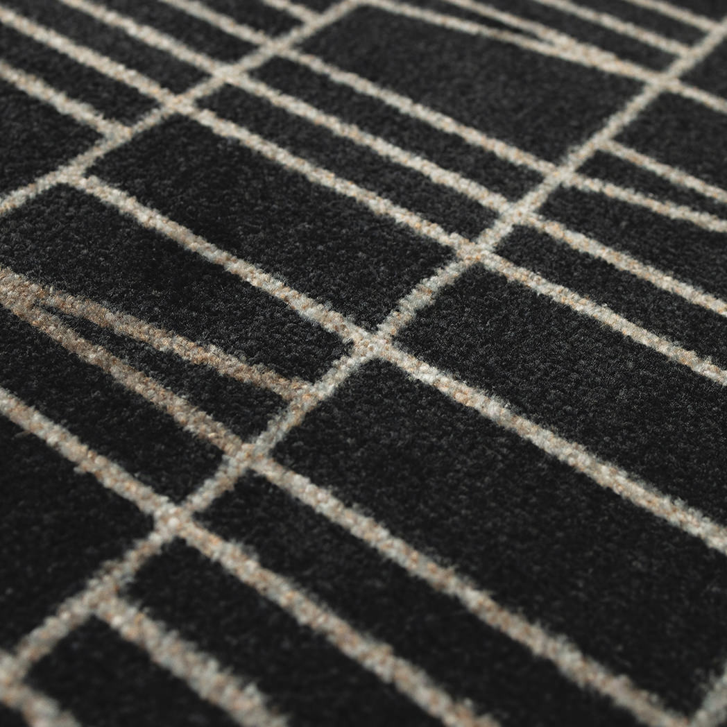 Close-up of the Maxcent Biblioteca area mat in charcoal, showing the soft carpeted surface with a modern beige line pattern over a deep charcoal background. The textured fibers provide a durable, comfortable feel underfoot.