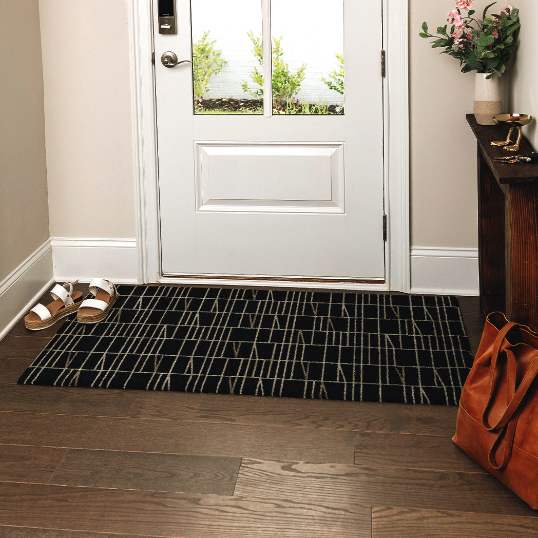 Maxcent Biblioteca area mat in Charcoal, medium size – placed at a front door on wood flooring with sandals and a bag nearby. The rug’s structured grid pattern adds a modern touch, while the non-slip backing keeps it stable in high-traffic entryways.
