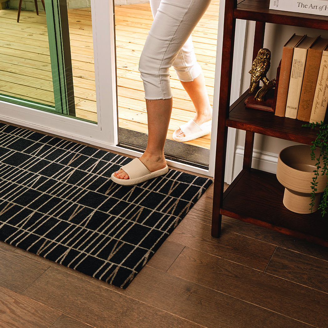 Maxcent Biblioteca runner rug in Charcoal – shown inside a sliding glass door on wood flooring, with a person stepping in from a deck. The black background and light tan linear pattern add a modern touch, while the non-slip backing helps the mat stay in place in transitional indoor-outdoor areas.