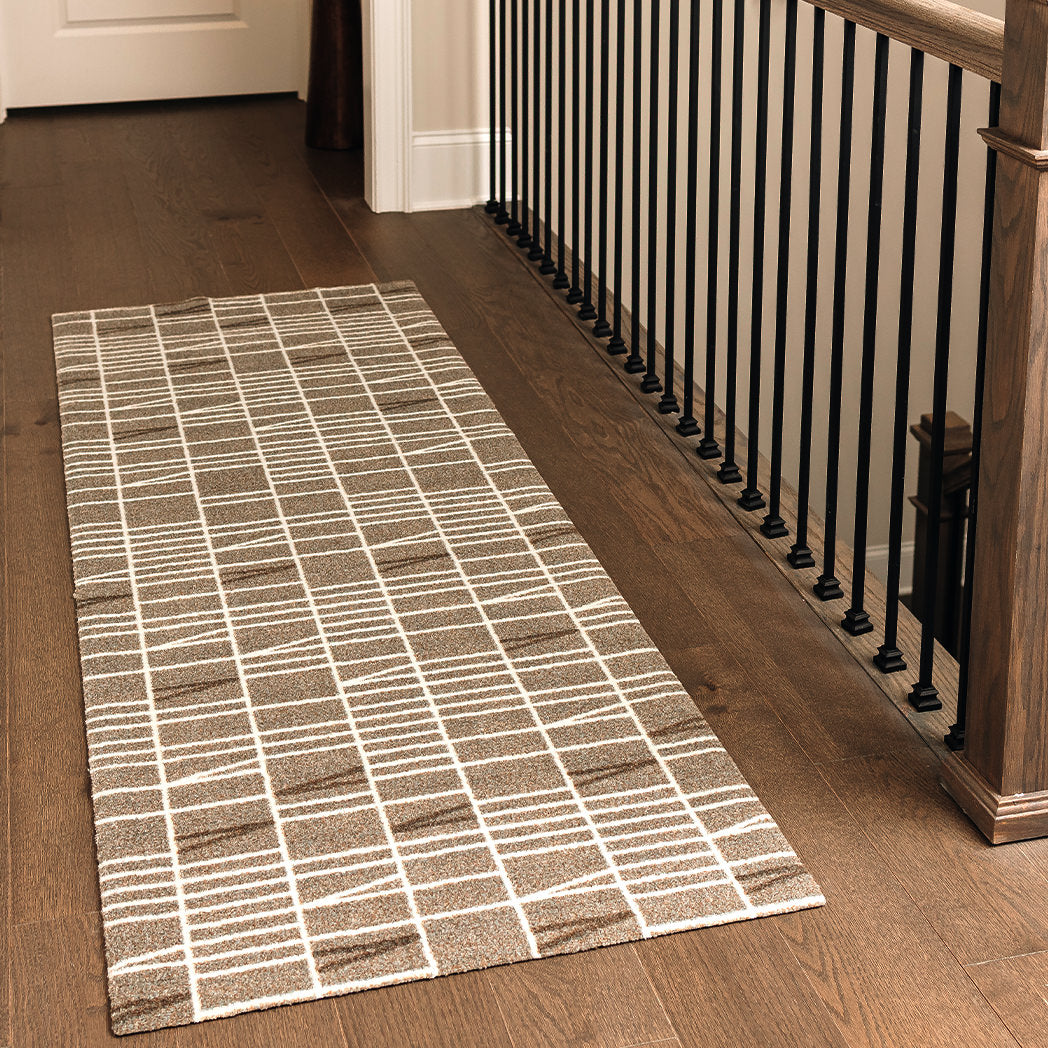 Maxcent Biblioteca runner rug in Light Greige – shown in a hallway with dark wood flooring and black stair railings. The rug features a soft beige-gray background with a white grid and angle pattern. Its non-slip backing and cushioned carpeted surface make it ideal for long, high-traffic areas like hallways or landings.