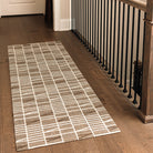 Maxcent Biblioteca runner rug in Light Greige – shown in a hallway with dark wood flooring and black stair railings. The rug features a soft beige-gray background with a white grid and angle pattern. Its non-slip backing and cushioned carpeted surface make it ideal for long, high-traffic areas like hallways or landings.