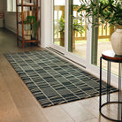 Maxcent Biblioteca runner rug in charcoal – a sleek, rectangular area mat with a modern grid-and-line pattern in beige on a dark charcoal background, shown in a bright entryway over hardwood flooring. Features a soft carpeted top and non-slip rubber backing for secure placement in high-traffic areas.