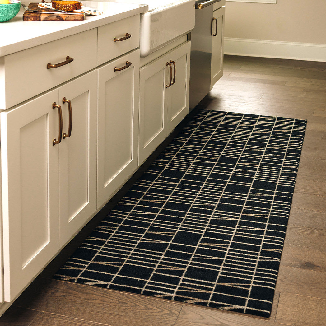 Maxcent Biblioteca runner mat in Charcoal – placed in a kitchen along white cabinetry and dark wood flooring. The rug features a black background with a light tan grid and angled line pattern. Its cushioned carpeted surface and non-slip rubber backing make it ideal for standing areas like sinks or prep spaces.