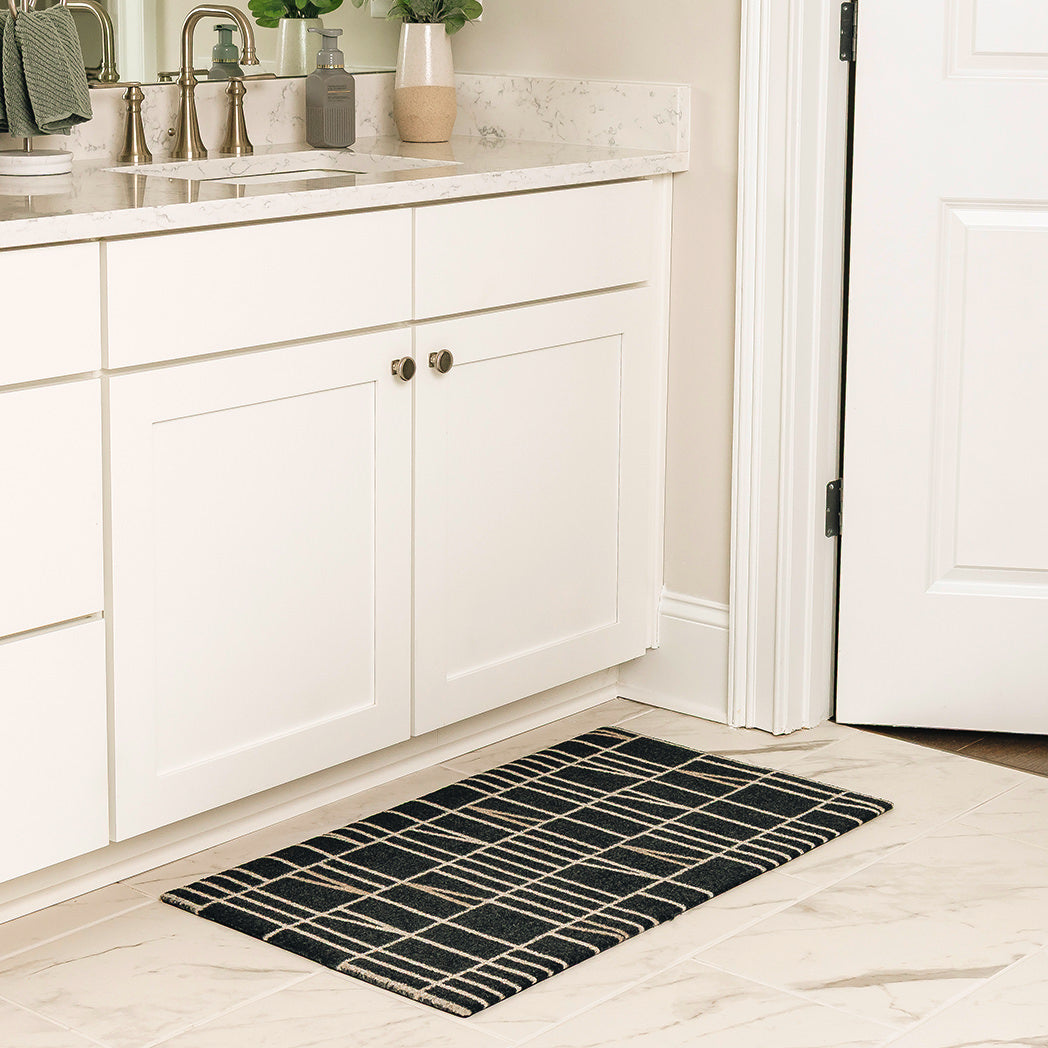 Maxcent Biblioteca area mat in charcoal, small size – shown in a bright bathroom in front of a white vanity. The low-profile rug features a clean grid pattern in beige over a dark charcoal base, with a non-slip rubber backing ideal for smooth tile floors.