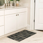 Maxcent Biblioteca area mat in charcoal, small size – shown in a bright bathroom in front of a white vanity. The low-profile rug features a clean grid pattern in beige over a dark charcoal base, with a non-slip rubber backing ideal for smooth tile floors.