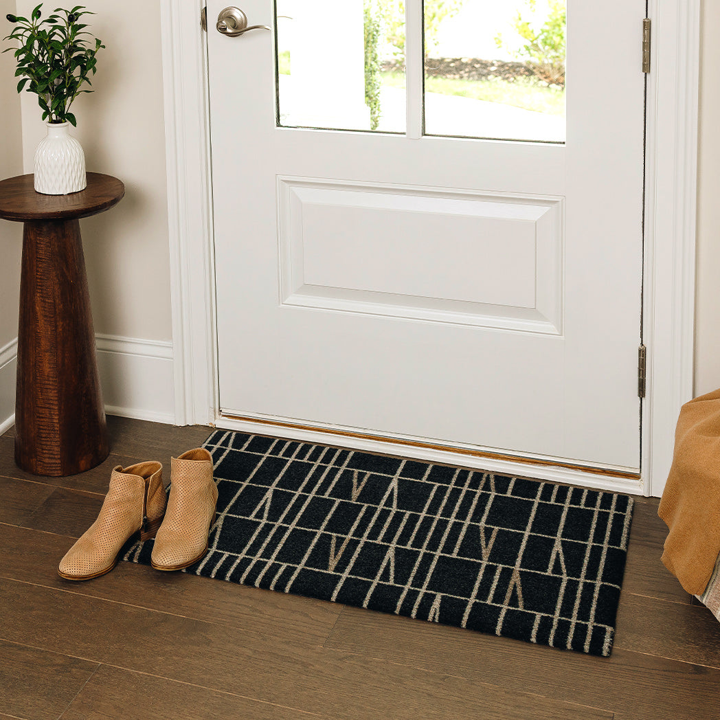 Maxcent Biblioteca mat in charcoal, small size – placed at a front door entryway on wood flooring. Features a contemporary beige line design over a dark charcoal background, with a non-slip rubber backing to keep the mat securely in place for everyday foot traffic.