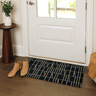 Maxcent Biblioteca mat in charcoal, small size – placed at a front door entryway on wood flooring. Features a contemporary beige line design over a dark charcoal background, with a non-slip rubber backing to keep the mat securely in place for everyday foot traffic.