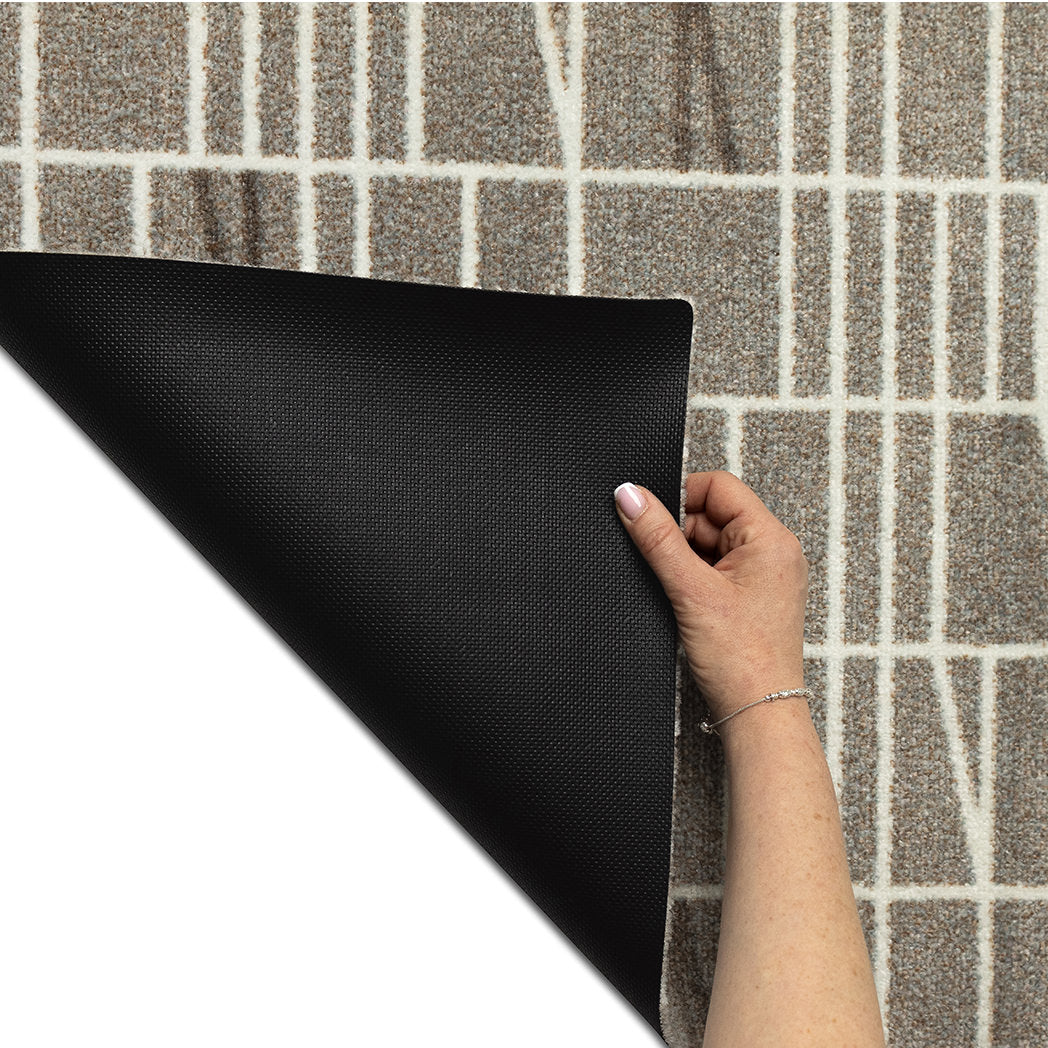 Hand lifting the corner of the Maxcent Biblioteca mat in Light Greige to show the textured, non-slip rubber backing. Designed for stability on hard floors, the mat features a modern grid pattern on a soft, carpeted surface.