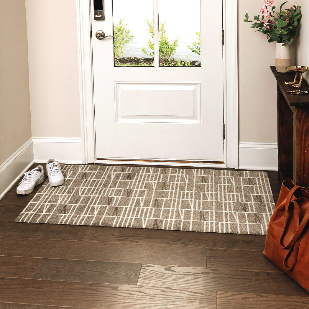 Maxcent Biblioteca area mat in Light Greige, medium size – shown in an entryway on dark wood flooring. The mat features a soft beige-gray background with off-white linear accents in a modern grid design. Its carpeted surface offers comfort, while the non-slip rubber backing keeps it secure in high-traffic areas.
