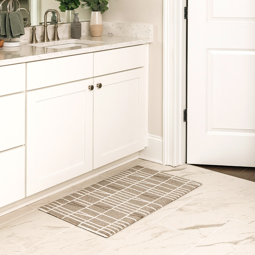 Maxcent Biblioteca area mat in Light Greige, small size – placed in front of a white bathroom vanity on marble tile flooring. The modern grid pattern in white on a light grey background and low-profile design offer a clean, contemporary look, while the non-slip backing helps keep the mat in place.