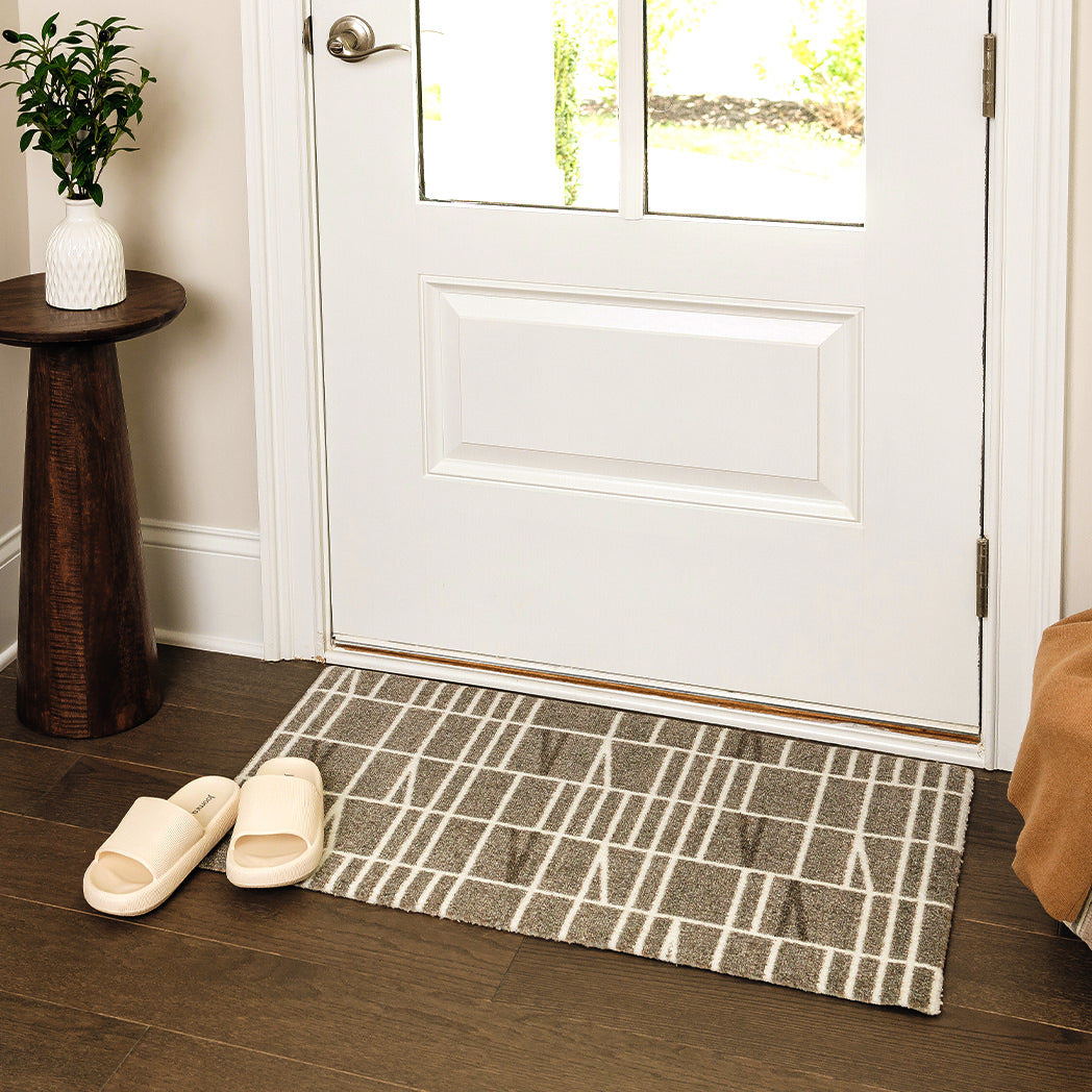 Maxcent Biblioteca area mat in Light Greige, small size – placed at a front door on wood flooring, shown with slippers nearby. The clean, grid-style pattern complements the entryway while the non-slip backing helps keep the mat firmly in place