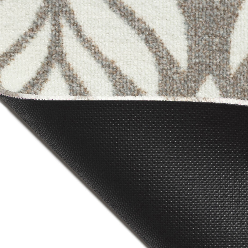 Corner of the Maxcent Botanica mat in a light colorway lifted to reveal the textured black rubber backing. The carpeted surface shows a soft brown and gray leaf pattern on a light cream background, offering both visual appeal and slip resistance on hard floors.