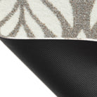 Corner of the Maxcent Botanica mat in a light colorway lifted to reveal the textured black rubber backing. The carpeted surface shows a soft brown and gray leaf pattern on a light cream background, offering both visual appeal and slip resistance on hard floors.