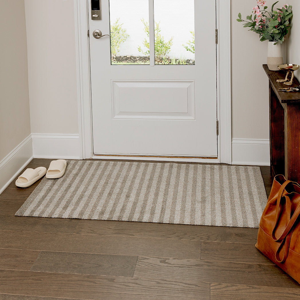 Maxcent Classic Stripe mat in medium size placed by a front entryway. The vertical striped design in soft beige and grey tones blends effortlessly with the neutral decor, offering a clean, welcoming touch while helping to trap dirt and moisture from shoes.