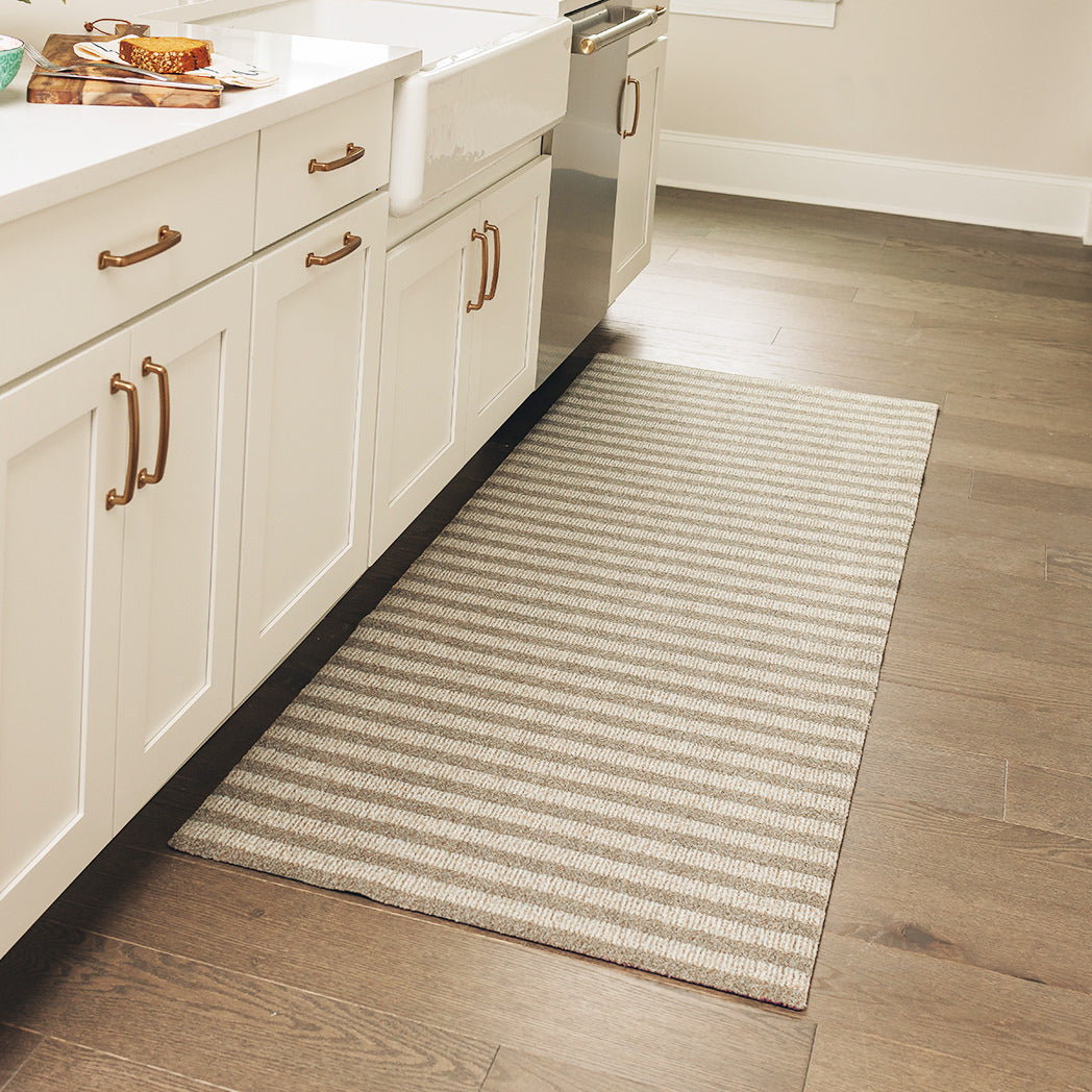 Maxcent Classic Stripe runner rug in a kitchen, featuring alternating beige and grey stripes. Positioned in front of the sink and cabinets, this long rug adds comfort and grip underfoot while protecting wood floors in a busy cooking area.