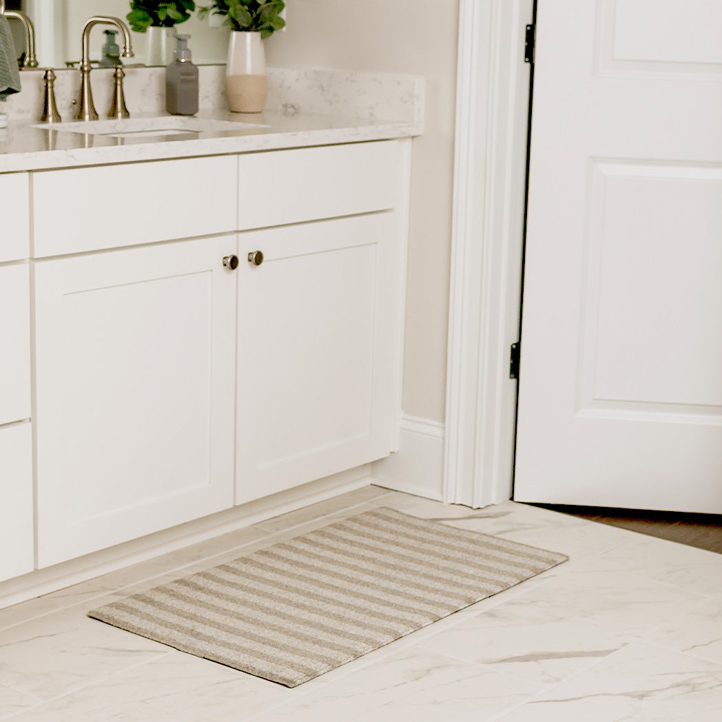 Maxcent Classic Stripe mat in a small size, placed in front of a bathroom vanity on white marble tile. The rug features a simple striped pattern in soft beige and grey tones, complementing the clean and modern space.