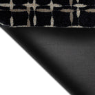 The corner of the Maxcent Heritage Tweed mat in Inkwell lifted to reveal the textured black rubber backing. The carpeted top displays a black background with a crosshatch pattern in light gray and tan, offering both grip and comfort for hard surface floors.