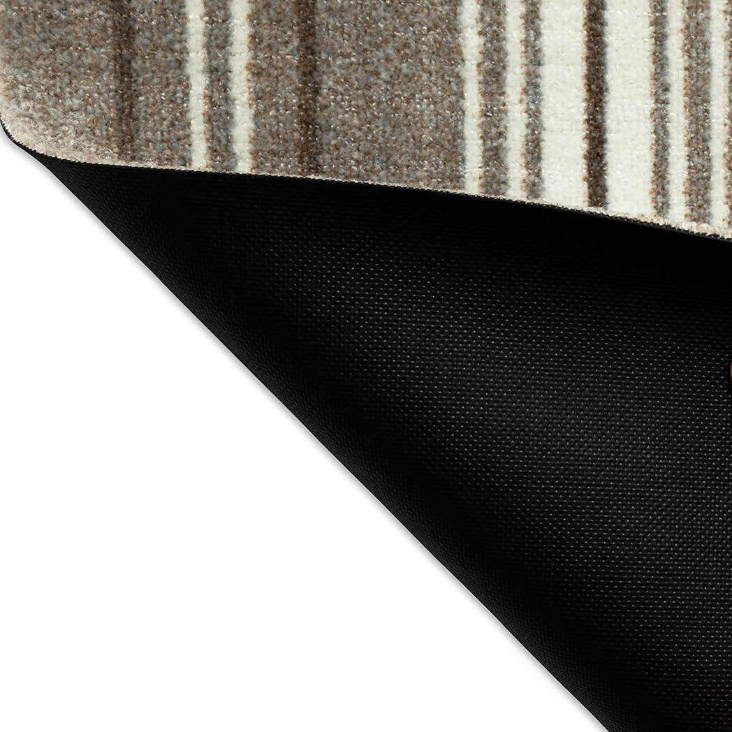 Corner of the Maxcent Ticking Stripes mat flipped over to reveal the durable black non-slip rubber backing, with the top side displaying warm beige, brown, and cream stripes."