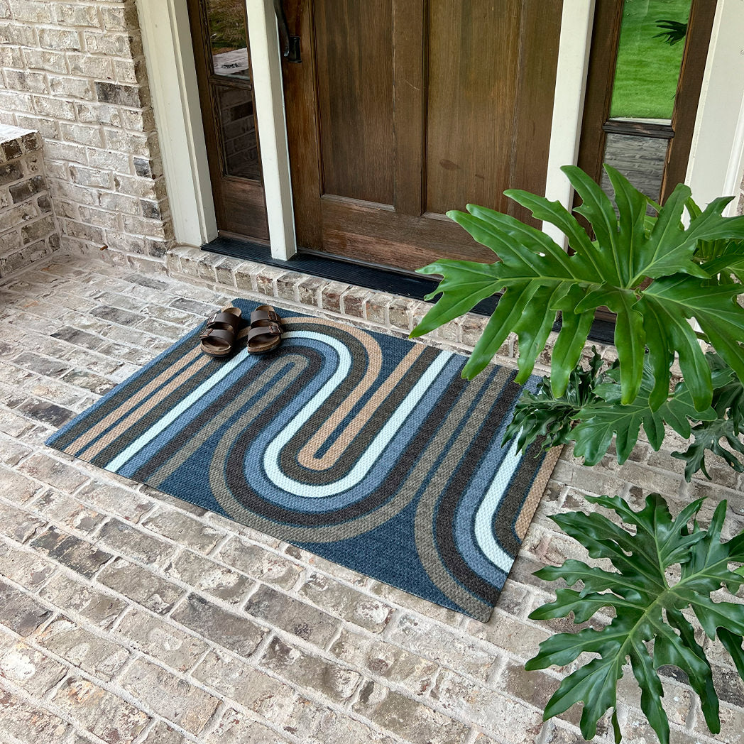 Decorative doormat with wavy pattern on a brick patio, shoes placed on it, and a plant to the right.