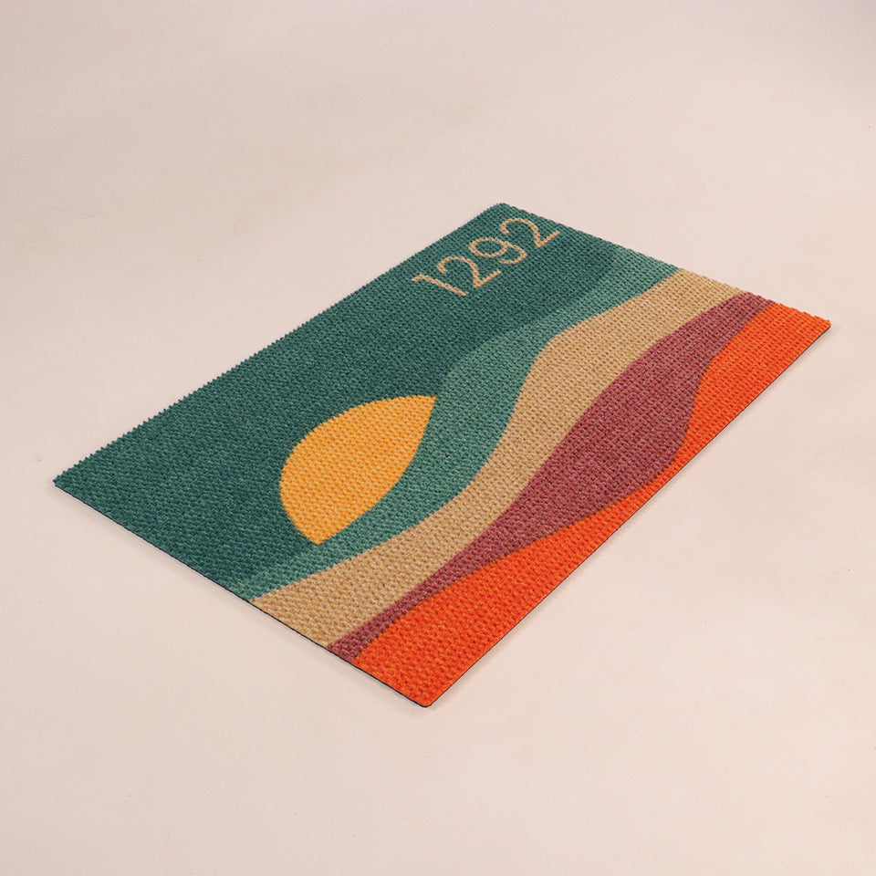 Angled left, single-door size image of Personal Sunset mat personalized with street address numbers and vibrant colors of aquas, coir, maroon, electric orange, and a yellow setting sun.