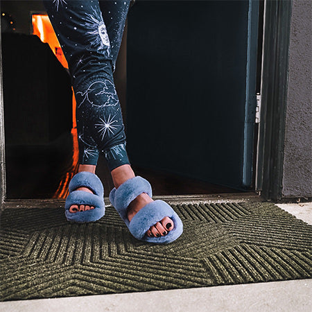 Fuzzy slippers standing on low profile WaterHog Lux outdoor doormat in olive green at front door of an apartment.