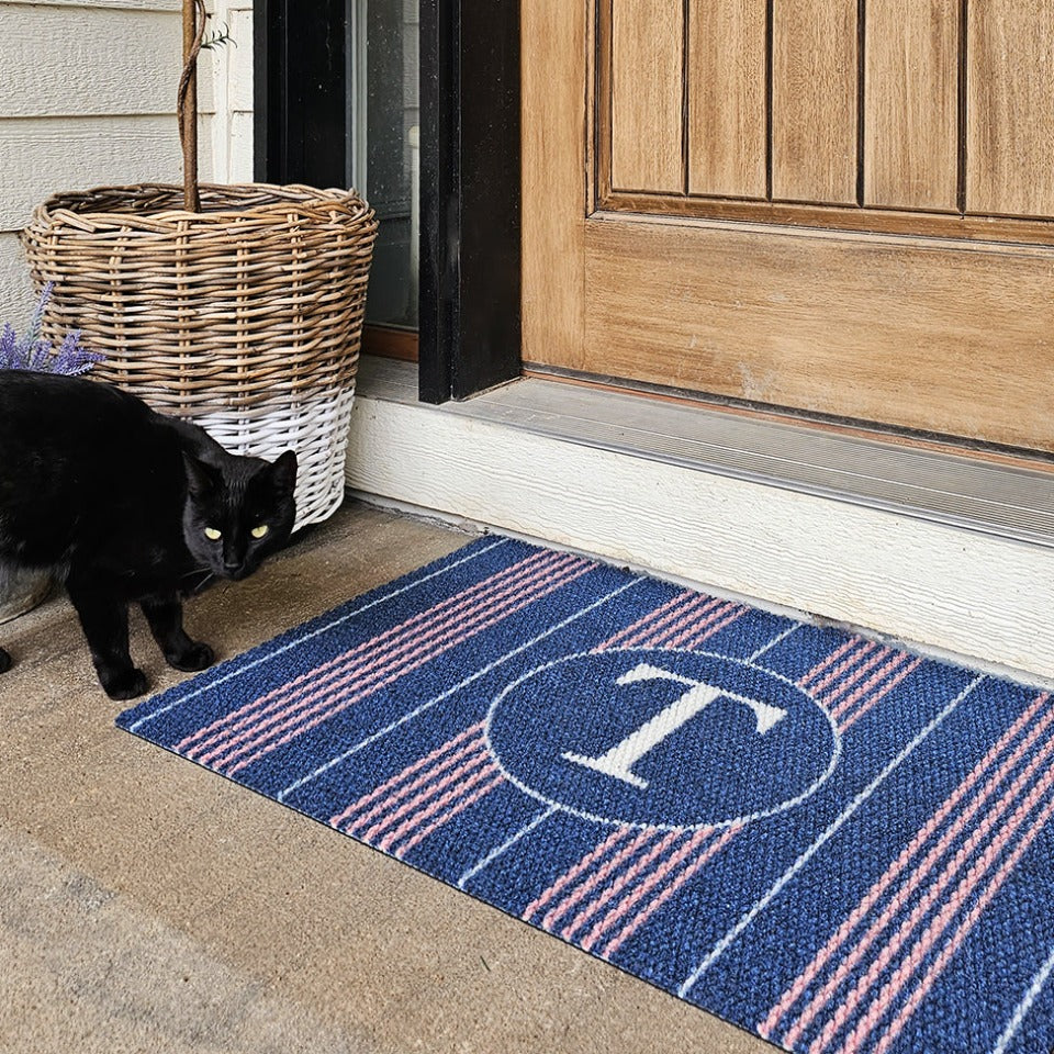 Neighburly Basic Stripe Monogrammed Decorative Doormat shown in blue and pink is placed just outside the front door with a black cat standing beside.