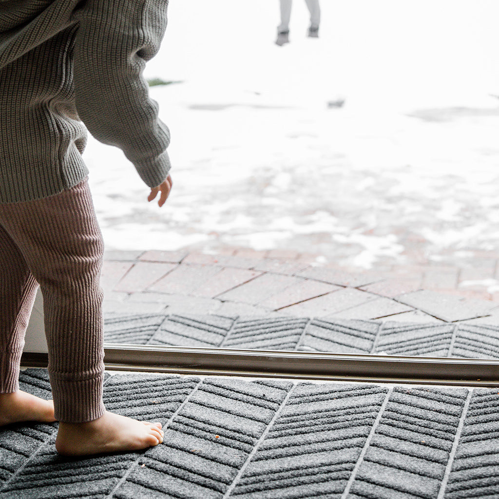 Small child standing on durable all-weather WaterHog outdoor doormat placed at sliding door to capture snow.