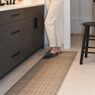 Happy Feet Brisbane Plaid runner mat in the color tan in  a bathroom with a woman in grey slippers standing on top of the mat