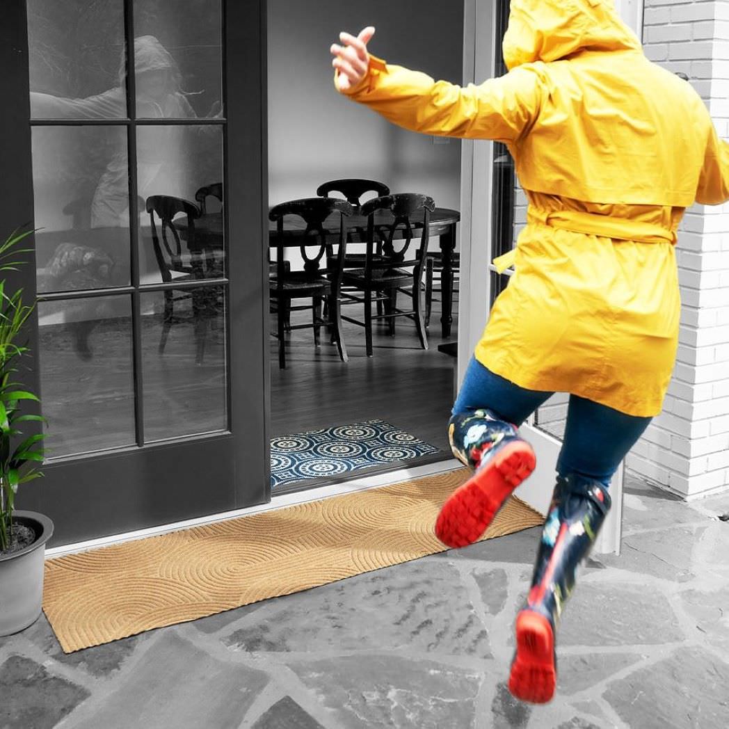 Durable WaterHog Luxe double door doormat placed in front of a set of double door glass doors outside with a young lady in a yellow rain coat and rain boots kicking her heels for fun.