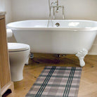 Low-profile and rubber backed Un-Rug shown in brown and tan plaid pattern is placed in front of a clawfoot bathtub.