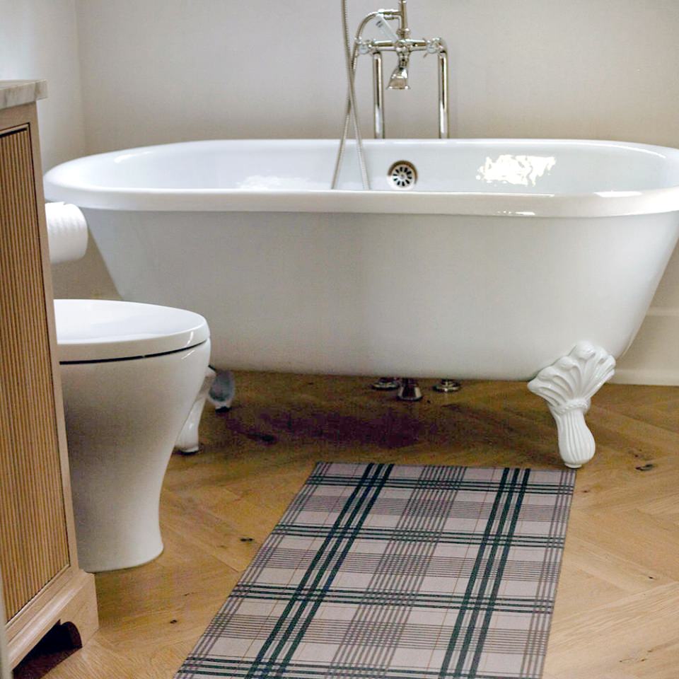 Low-profile and rubber backed Un-Rug shown in brown and tan plaid pattern is placed in front of a clawfoot bathtub.