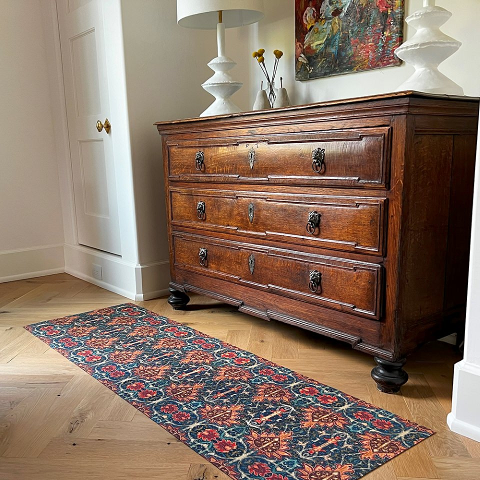 This Un-Rug shown in front of a bedroom dresser is a colorful vibrant Persian style rug with burnt orange, navy, teal, greens, yellows, blues in a repeating ornate pattern on a washable indoor floor mat.