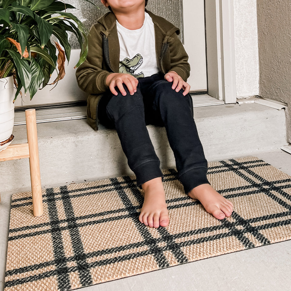 Young boy sitting on a doorstep with his bare feet on a Neighburly Classic Plaid single door mat that coir with black lines.