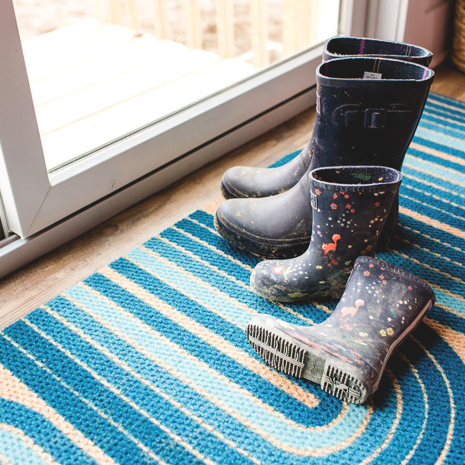 A modern luxury doormat, the Neighburly Retro Vibes is placed at a set of glass sliding doors with dirty rain boots on top.