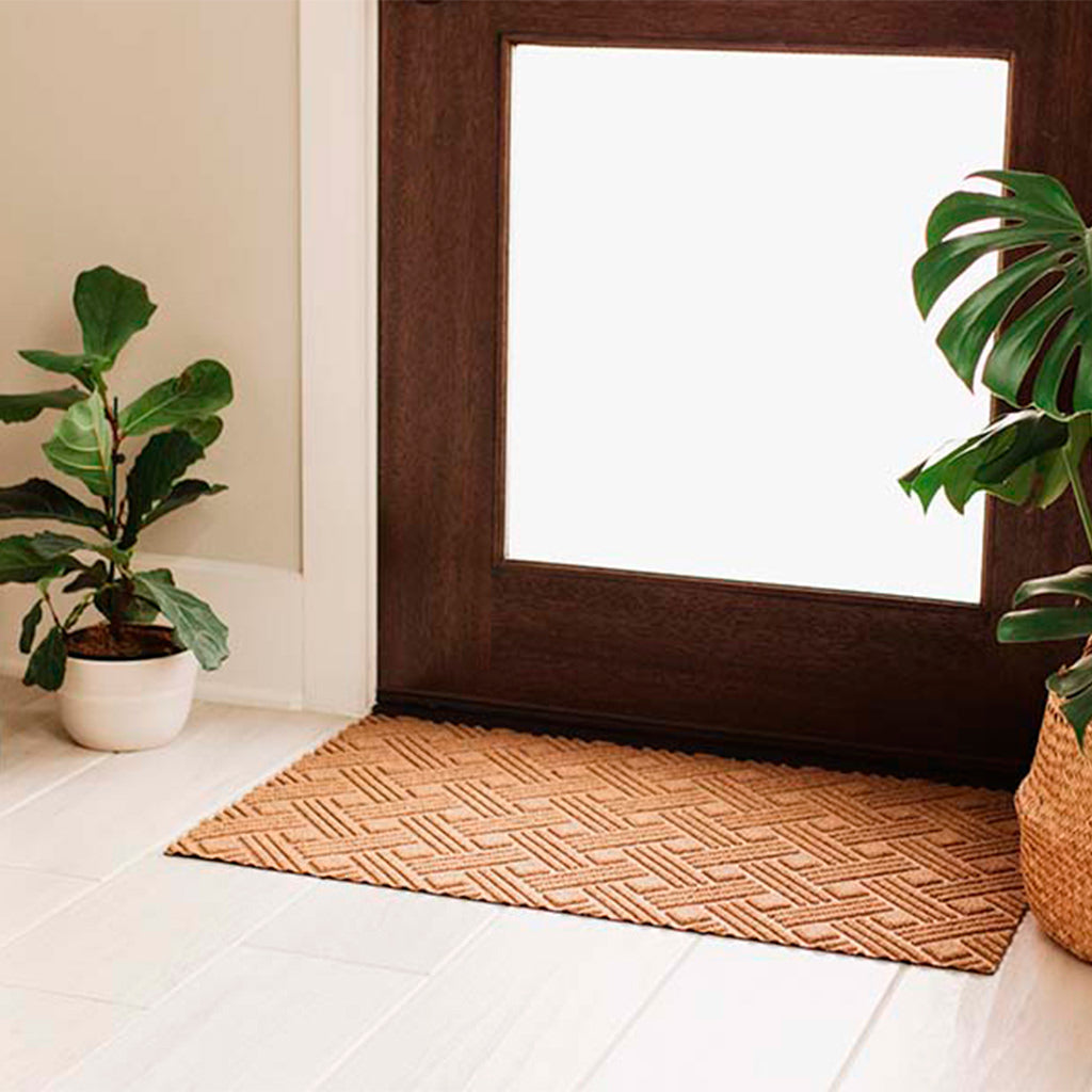 Wheat colored WaterHog Luxe Classic Thatch doormats inside a closed door surrounded by two plants.