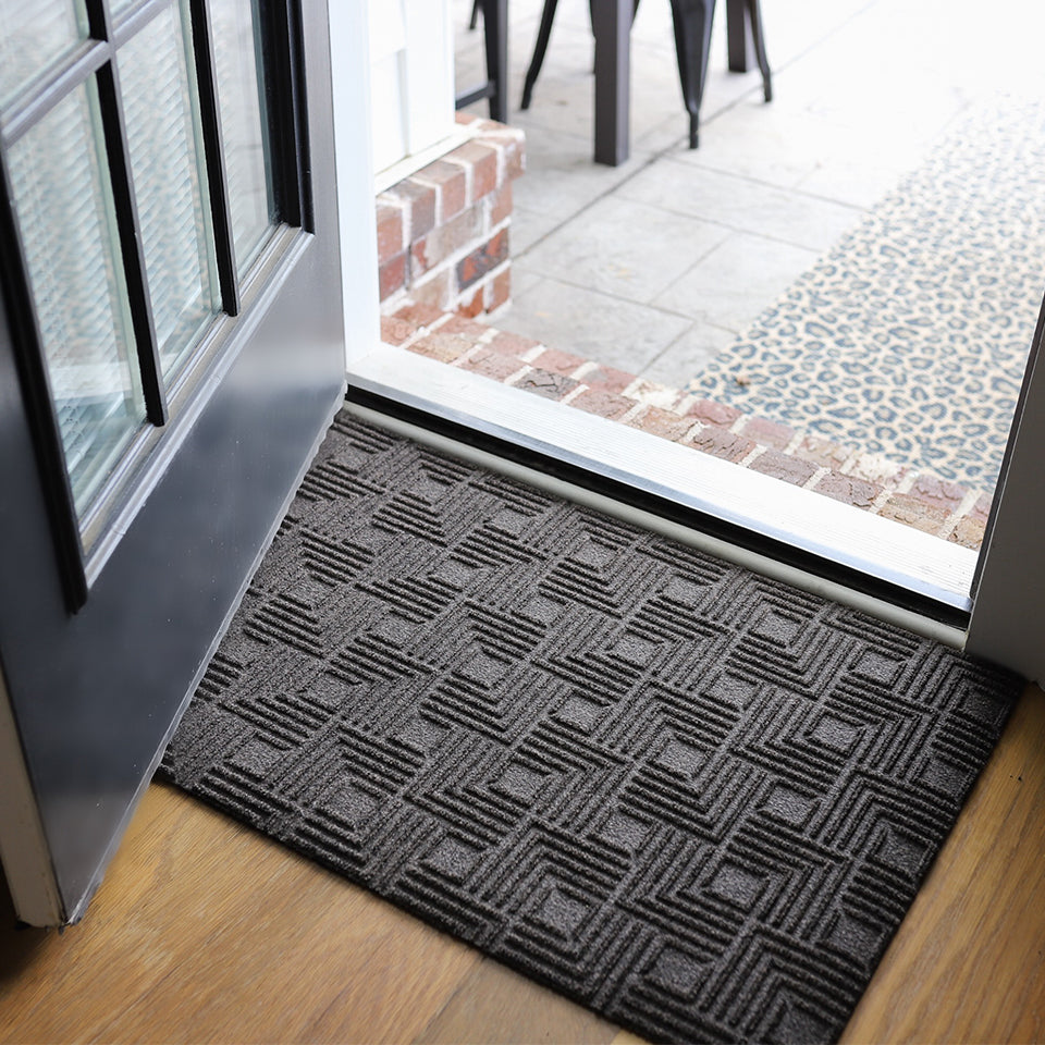 An all-weather Labyrinth doormat at the entrance of a home, below an opened door. In the color graphite.