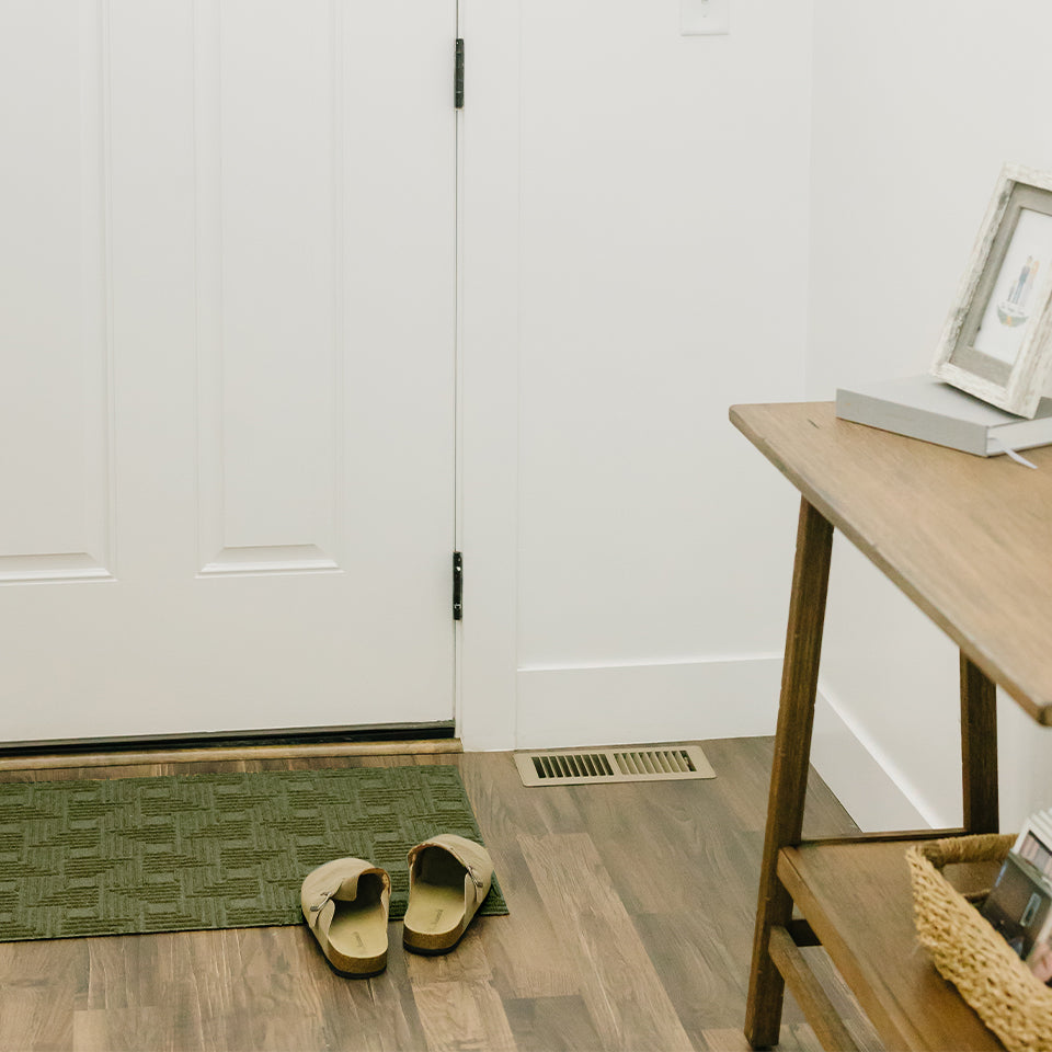 A WaterHog Luxe Labyrinth doormat in olive lying in front of a closed door of a home.