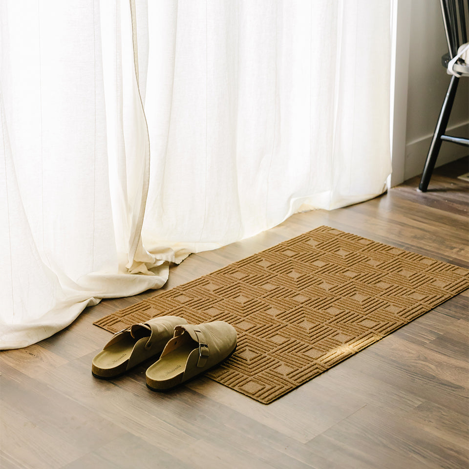 Dreamy picture of a Labyrinth mat next to creamy curtains casting soft light and a pair of slippers half on the wheat-colored mat.