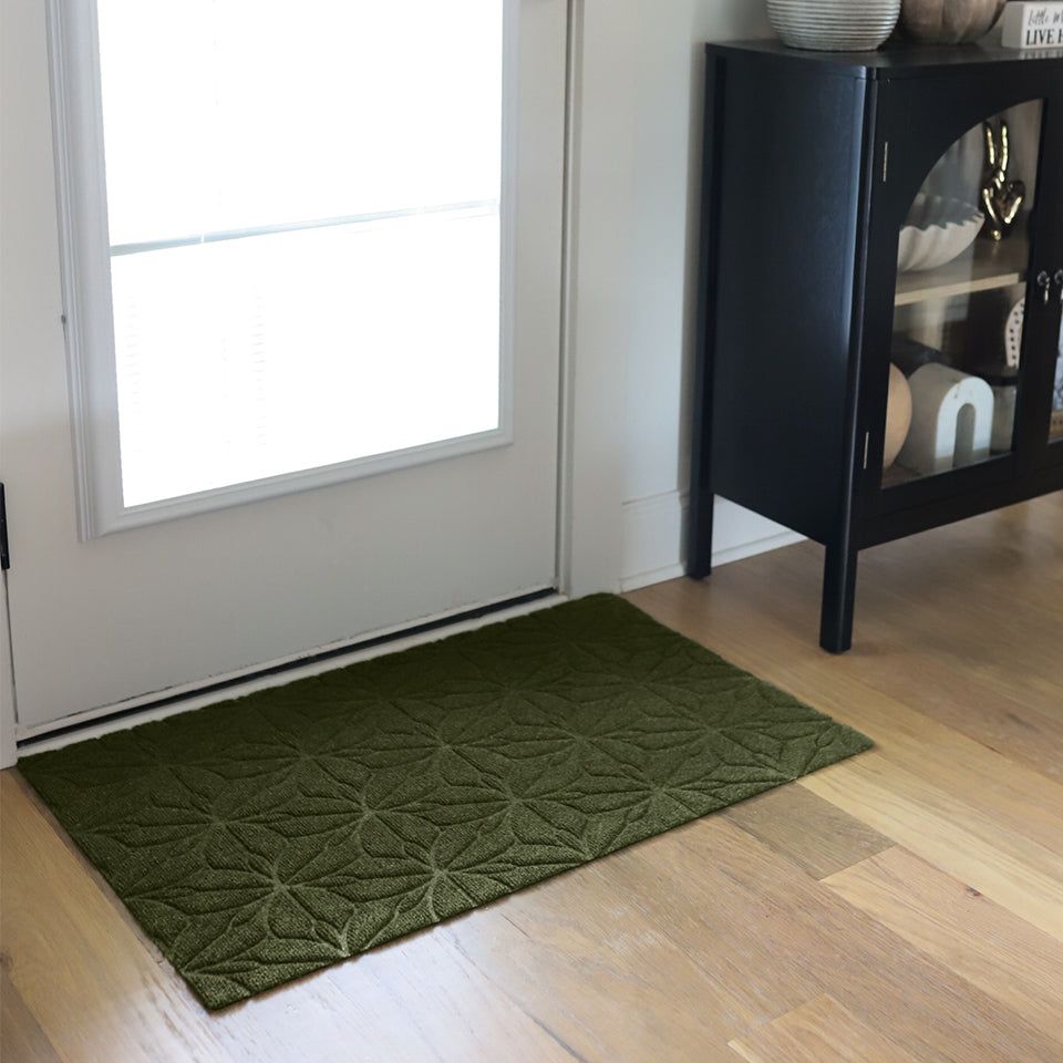 An angled image of the olive WaterHog Luxe Magnolia mat at the door with light shining through the glass, creating contrast to show off the bi-level surface and symmetrical pattern.